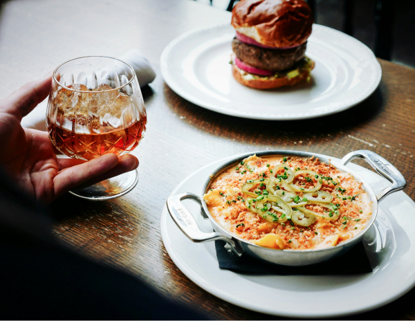 Image of someone holding a glass of whiskey over a table with a burger and a dip